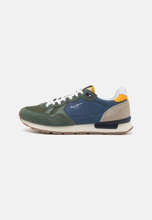 Sneakers in suede verde e blu navy con lacci bianchi, caratterizzati da un tallone beige e dettagli gialli. Si può notare il tessuto testurizzato e una suola in gomma.