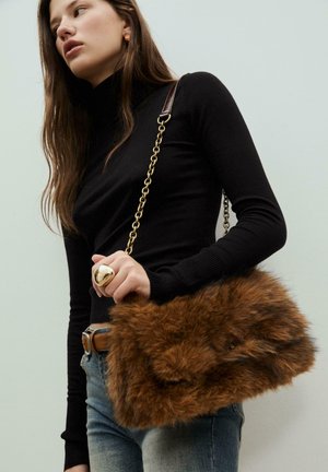 Braune Kunstfell-Handtasche mit einer Kettenriemen, rechteckiger Form und strukturierten Oberfläche. Das Model trägt einen schwarzen Rollkragenpullover und Jeans.