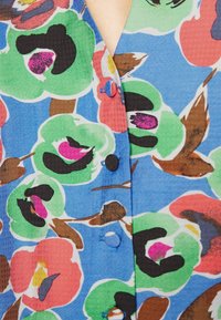 Vêtement à boutons fleuri avec un fond bleu, mettant en avant de grandes fleurs vertes, roses et rouges, des accents marron et des détails noirs.