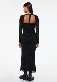 Eine Frau, die sich abwendet, trägt ein langes schwarzes Kleid aus floralem Spitzenmaterial mit offenen Rücken, das am Hals gebunden ist, und klobige schwarze Stiefel.