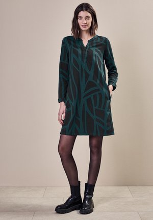 Abito verde e nero a fantasia con maniche lunghe, scollatura a V e tasche laterali, abbinato a stivaletti neri e collant trasparenti.