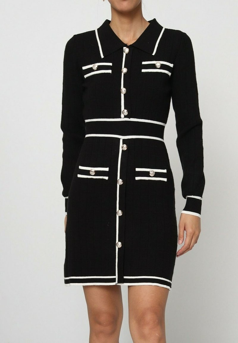 Robe tricotée noire avec des rayures blanches, à manches longues et col. Dispose de quatre poches et de boutons ronds devant. Tissu texturé.