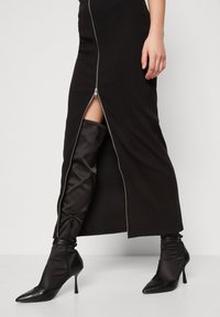 Schwarzes geripptes Kleid mit einem Reißverschluss vorne und seitlichem Schlitz, kombiniert mit knielangen schwarzen Satin-Stiefeln. Die Stiefel mit Absatz haben eine spitze Zehenform.