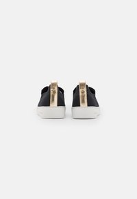 Ein Paar schwarze Slip-On-Sneaker mit weißen Sohlen und goldenen Fersenklappen, die ein kreisförmiges Logo tragen. Von hinten auf einem weißen Hintergrund gezeigt.