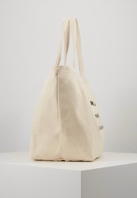 Beige canvas tote bag with two handles, standing upright on a white pedestal against a plain gray background.