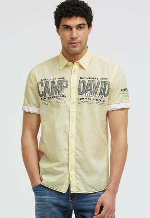 Mann trägt gelbes kurzärmeliges Hemd mit "Camp David" Text, steht mit einer Hand in der Tasche vor einfarbigem Hintergrund.