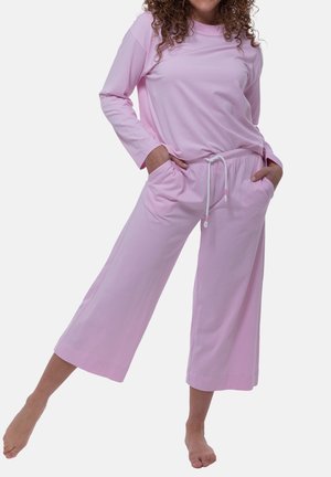 Vrouw draagt lichtroze top met lange mouwen en losse korte broek, staat blootsvoets met handen in de zakken en één been opgetild.