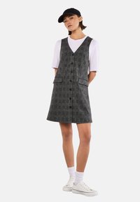 Vestido de cuadros gris con escote en V, botones en el frente y dos bolsillos frontales, combinado con una camiseta blanca y una gorra negra. Zapatillas blancas completan el conjunto.