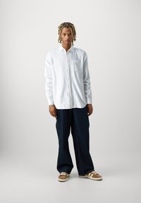 Camisa blanca de manga larga con botones y bolsillo en el pecho, combinada con vaqueros oscuros de corte suelto y zapatillas marrones con rayas blancas.