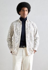 Camisa de manga larga en color crema con un patrón de paisley azul, cierre con botones en la parte delantera y tejido suave; llevada sobre un cuello de tortuga oscuro y pantalones de color claro.