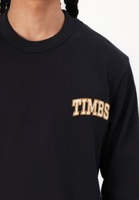 Zwarte lange mouwen shirt met een ronde halslijn en een gouden geborduurd "TIMBS" logo op de linkerkant van de borst. Zachte katoen stof.