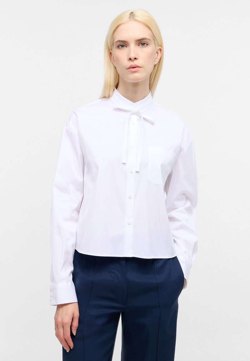 Witte katoenen shirt met een Mandarin kraag, strikdetail en lange mouwen. Beschikt over een enkele borstzak en knoopsluiting.