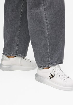 Balti sportiniai bateliai su blizgiu sidabriniu padu, turintys grafikos akcentą ir raištelių dizainą, derinami su pilka džinsine kelnėmis.