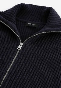 Marine gebreide ritssluiting trui met een geribbelde textuur, hoge kraag en zilveren ritsdetail. Etiket leest "NEXT."