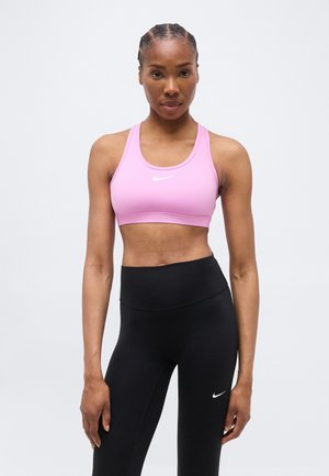 Donna in piedi di fronte, indossa un reggiseno sportivo rosa e leggings neri a vita alta con un logo Nike bianco su entrambi.