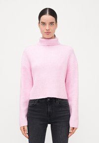 Pulover roz deschis cu nervuri, cu guler înalt și mâneci lungi, având un design scurt. Purat cu blugi negri.
