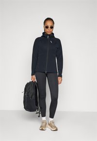 Schwarze Zip-Jacke mit Kapuze, schwarze Leggings, beige Sneakers und einen schwarzen Rucksack. Das Outfit hat ein schlankes, sportliches Design.