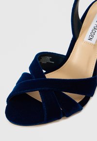 Tumši zili samta augstpapēžu sandales ar krustenisku siksnu dizainu, atvērtu purngalu un gludu bēša iekšējo zoli. Iekļauj izturīgu zoli un minimālistisku stilu.