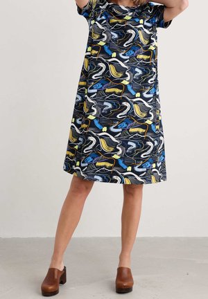 Femme portant une robe à manches courtes, longueur genou avec un motif abstrait coloré et des sabots marron à talons sur un sol gris clair.