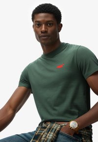 Mand iført grøn Superdry t-shirt, ternet skjorte knyttet om livet, blå jeans og guldur, poserer mod en hvid baggrund.
