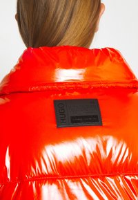 Doudoune rouge brillante avec un col montant, une étiquette de marque noire au dos et une texture matelassée. Surface lisse avec une finition brillante.