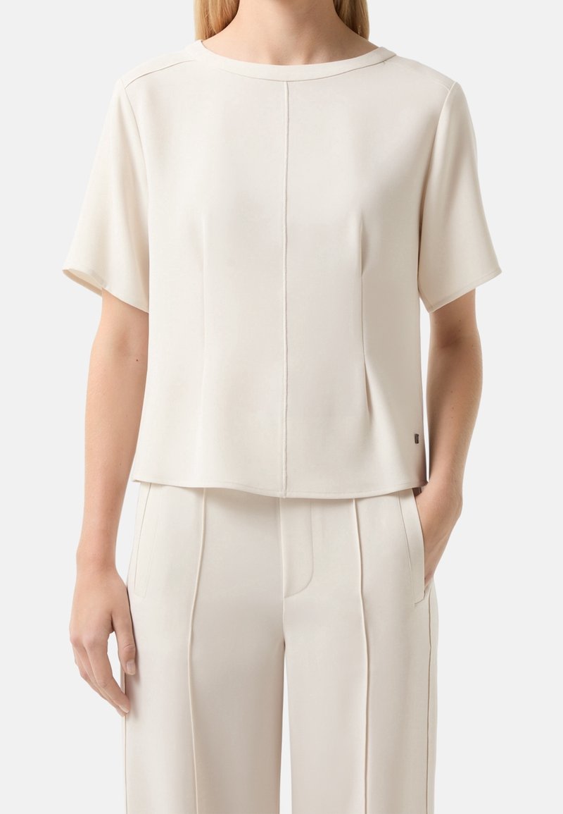 Femme portant un haut à manches courtes crème et un pantalon crème assorti avec des détails de couture verticale, une main dans la poche, sur un fond uni.