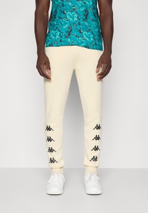 Un homme porte un jogging crème avec des logos Kappa noirs sur les jambes inférieures, une chemise camouflage bleu-vert et des baskets blanches, se tenant devant un fond blanc.