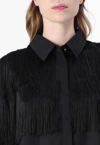 Camisa preta com um design em franjas na parte da frente e nos ombros, com colarinho clássico e fecho de botão. Textura suave.