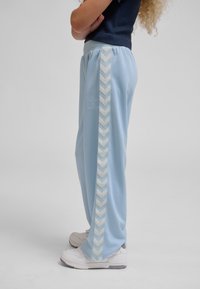 Kvinde står med armene krydset iført lyseblå træningsbukser med hvide chevronstriber og hvide sneakers mod ensfarvet baggrund.