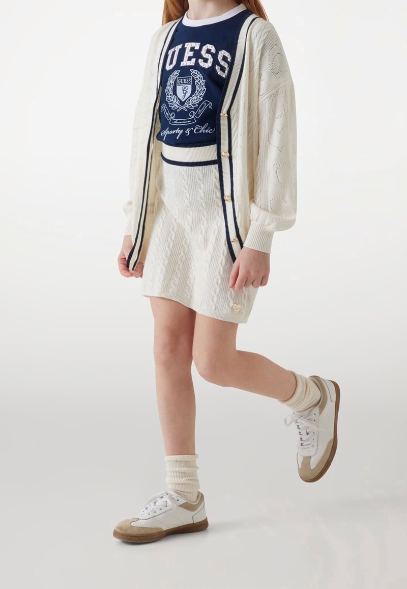 Enfant marchant, portant une jupe torsadée crème et un cardigan, un t-shirt graphique bleu marine, des baskets blanches avec des touches beiges et des chaussettes crème.