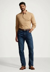 Beige langermet polotrøye, mørk denimjeans, brun skinnbelte, og brune sko. Mann står selvsikkert mot en nøytral bakgrunn.