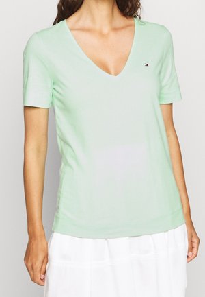 Woman wearing a light green V-neck short sleeve top with a small logo on the chest, paired with a white skirt.