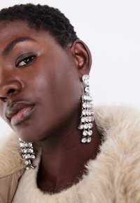 Sølv dråpeøreringer med flere klare rhinestones, med et kaskadedesign og teksturerte overflater, satt mot en nøytral bakgrunn.
