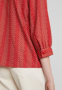 Rød bluse med fiskebensmønster, der har puffede ærmer, elastiske manchetter og en enkelt knaplukning. Kombineret med beige bukser.