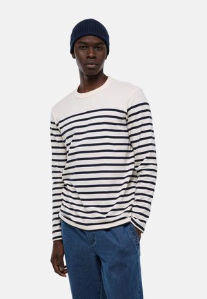 Homme portant un bonnet bleu marine, une chemise blanche à manches longues avec des rayures horizontales bleu marine, et un jean bleu, debout avec une main dans la poche.