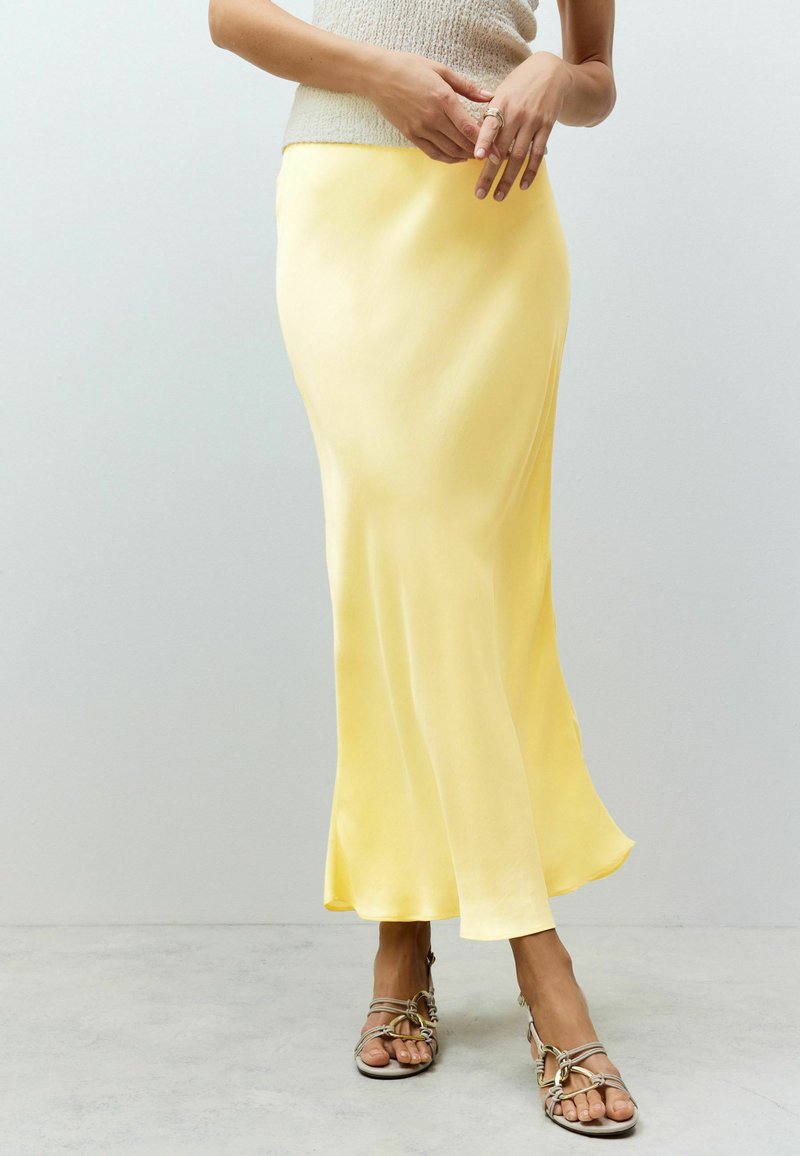 Jupe maxi en satin jaune avec une silhouette fluides, une texture lisse et un léger éclat, associée à des sandales à lanières neutres.