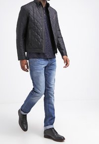 Svart quiltad jacka med dragkedja och tryckknappslåsg, kombinerad med en mörk polo, ljusblå jeans och svarta läderskor.