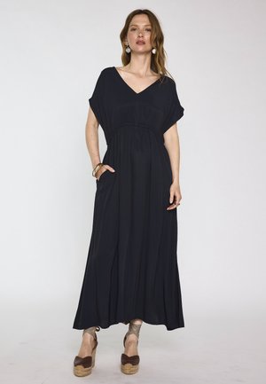 Femme portant une robe longue noire à décolleté en V avec des manches courtes, debout avec une main dans la poche, associée à des sandales à talons compensés marron et des bijoux.