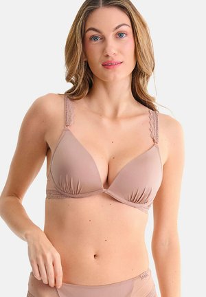 Femme à la peau claire et aux longs cheveux portant un soutien-gorge mauve clair avec des détails en dentelle, posant devant un fond blanc uni.