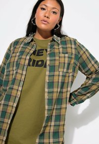 Groen en gele geruite overhemd met een knoopsluiting, voorzien van een met edelstenen versierde kraag en een borstzak, gelaagd over een olijfgroen T-shirt met zwarte tekst.