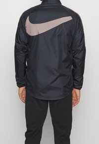 Marinblå vindjacka med långa ärmar och en stor brun Nike swoosh på baksidan, i en mjuk och slät textur.