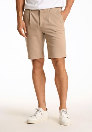 Beige tailored shorts with pleats, a flat front, and side pockets. The fabric looks smooth with a lightweight finish. Paired with white sneakers.