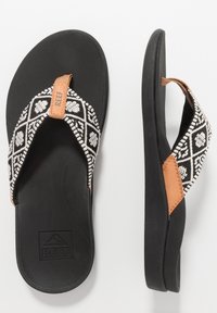Zwarte slippers met een gestreepte stoffen band in zwart en wit, met een bruine leren accent en gestructureerde zwarte rubberen zolen.