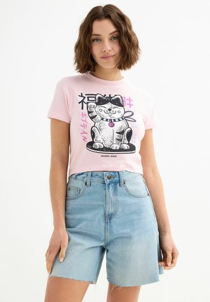 Camiseta rosa de manga corta con un gráfico en blanco y negro de un gato saludando, acompañado de texto en japonés, combinado con pantalones cortos de mezclilla azules de talle alto.
