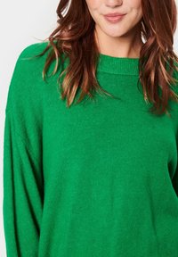 Vihreä neulepaita, jossa on leveä kaula-aukko, rento leikkaus ja teksturoitu kangas. Pitkät hihat kapenevat ranteiden kohdalla.