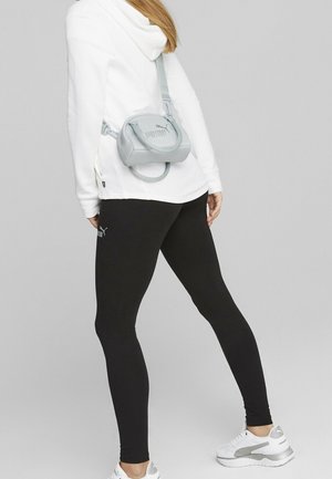 Personne portant des leggings noirs Puma, un sweat à capuche blanc et des baskets blanches, avec un petit sac banane gris Puma porté dans le dos.