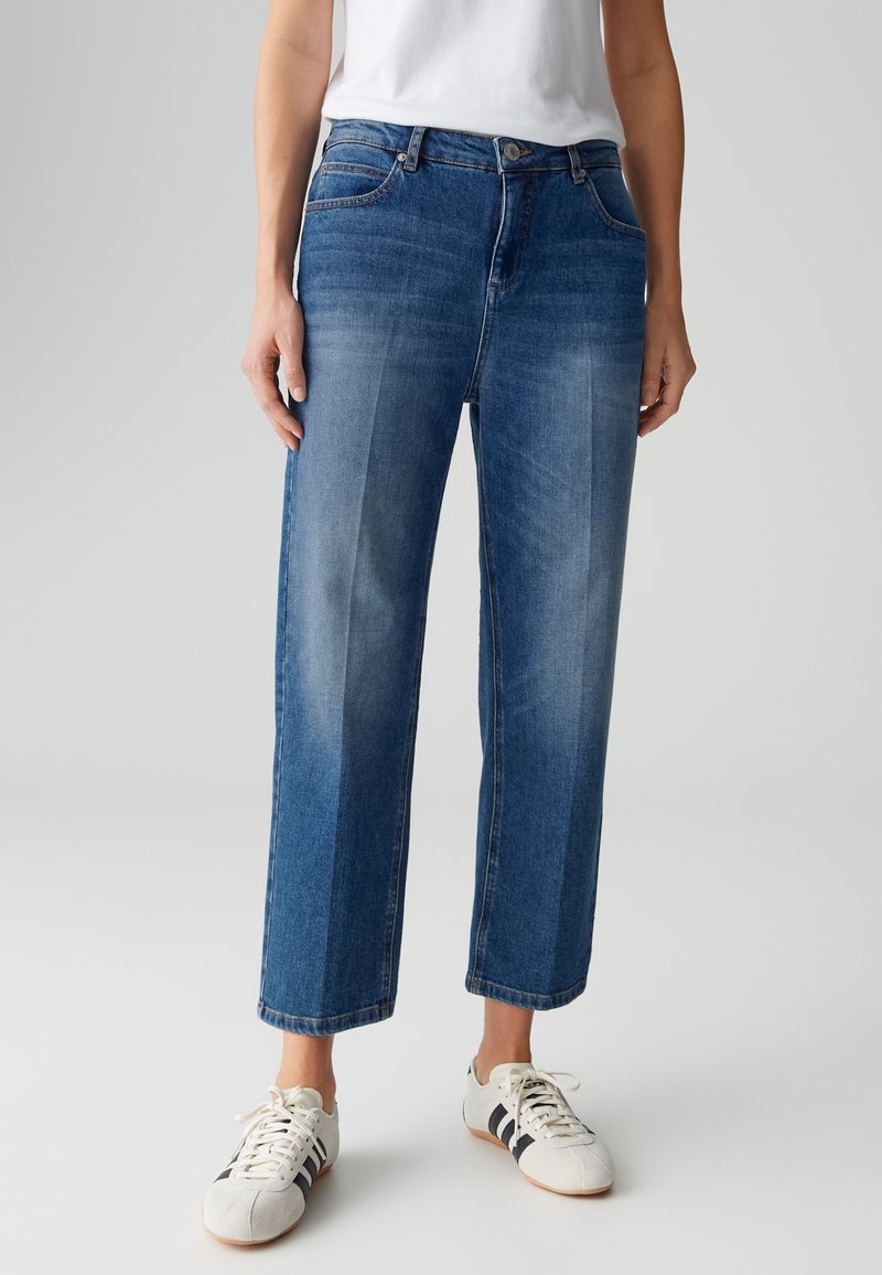 Personne portant un jean bleu taille mi-haute, coupe droite, avec des panneaux verticaux décolorés et des baskets blanches avec des bandes noires.