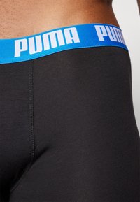 Juodos tvirtos kelnaitės su kontrastinga mėlyna juostele, ant kurios užrašyta "PUMA" balta spalva. Lygus paviršius ir tampri medžiaga.