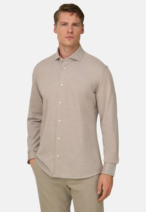 Homme portant une chemise beige à manches longues boutonnée et un pantalon assorti, debout avec une main dans la poche devant un fond uni.