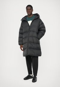 Moški nosi dolgo črno puffer jakno s kapuco, temne hlače, črne superge, stoji z eno roko v žepu ob beli ozadju.